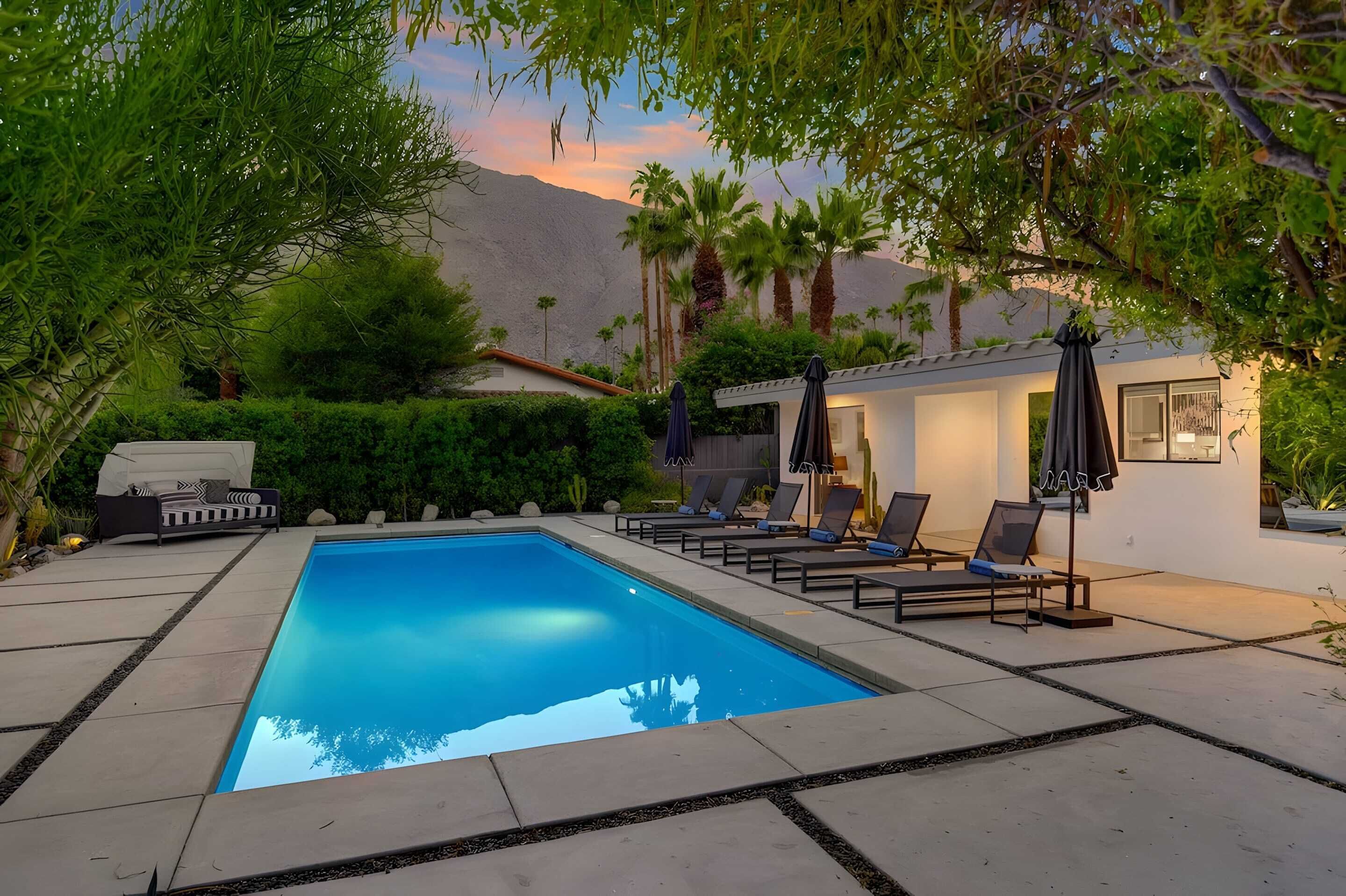 A rectangular swimming pool with lounge chairs and umbrellas, surrounded by greenery and a mountain backdrop.
