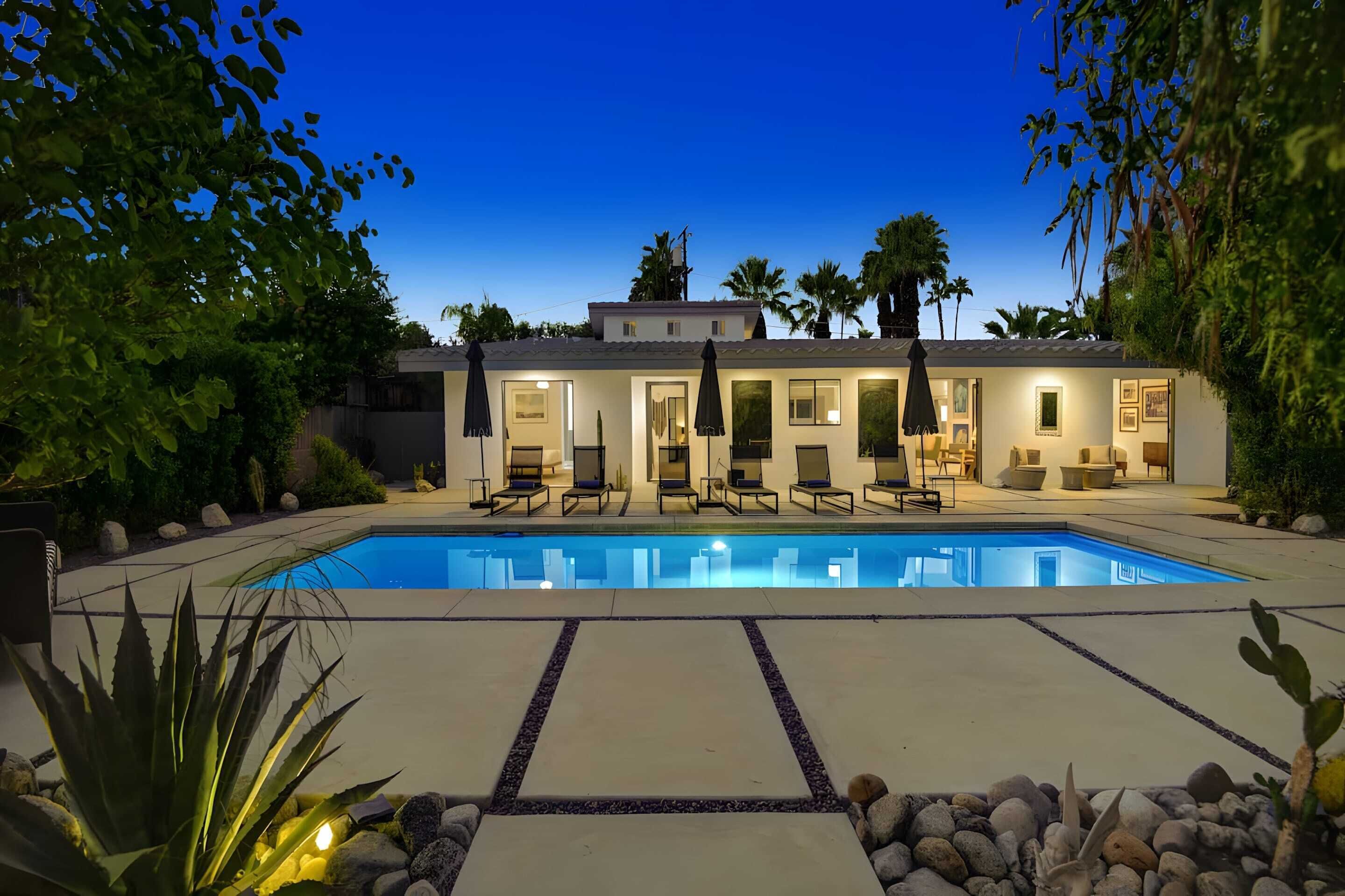 A poolside area with a swimming pool, lounge chairs, and a well-lit building with multiple doors and windows.