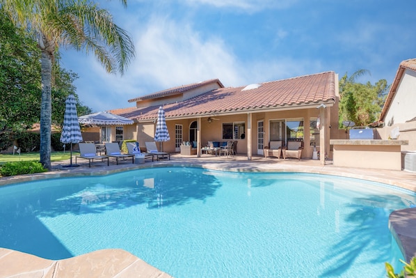 A swimming pool with lounge chairs and umbrellas, a covered patio area, and a house with a tiled roof.