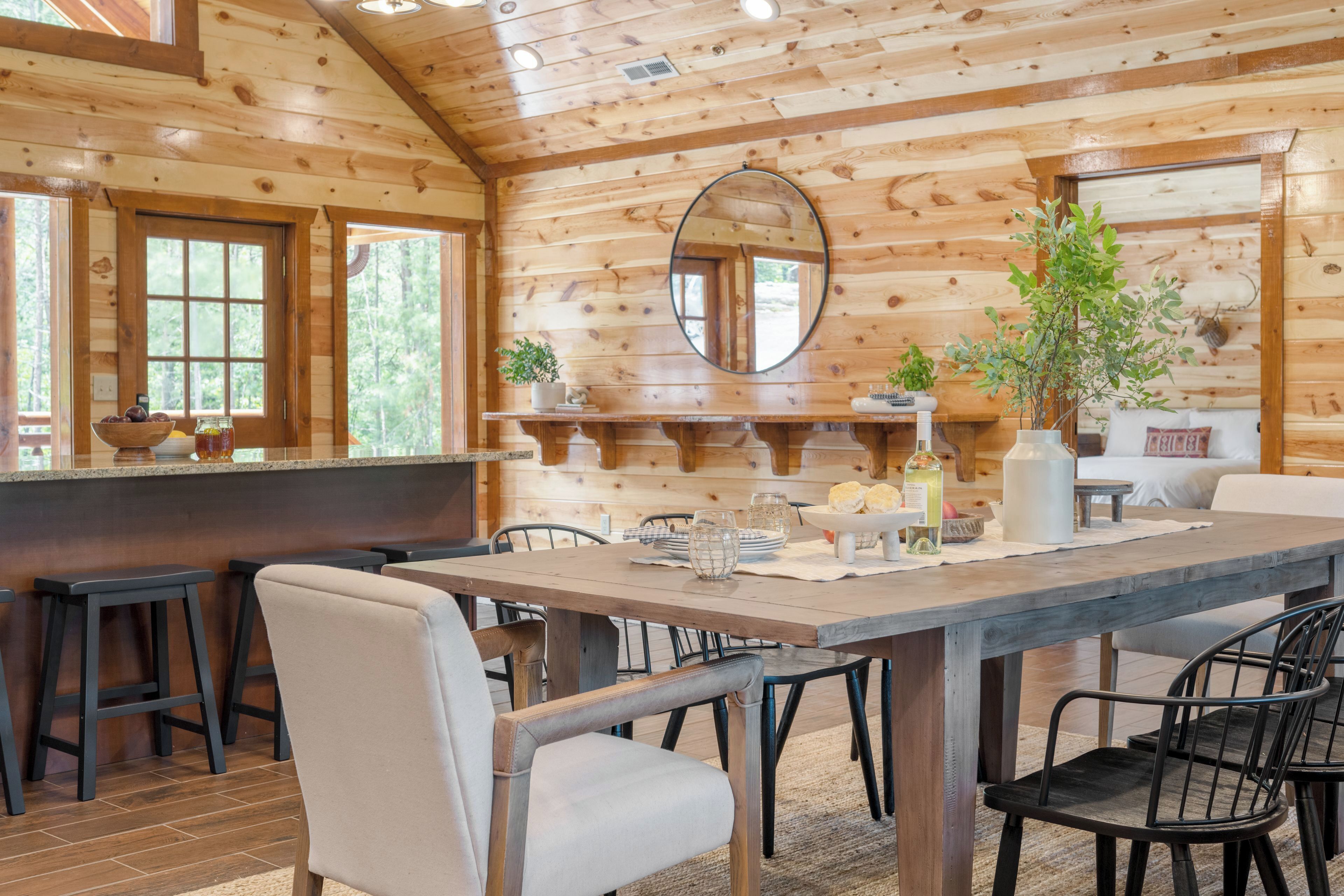 A wooden cabin interior with a dining table, chairs, a bar area, and a large mirror on the wall.