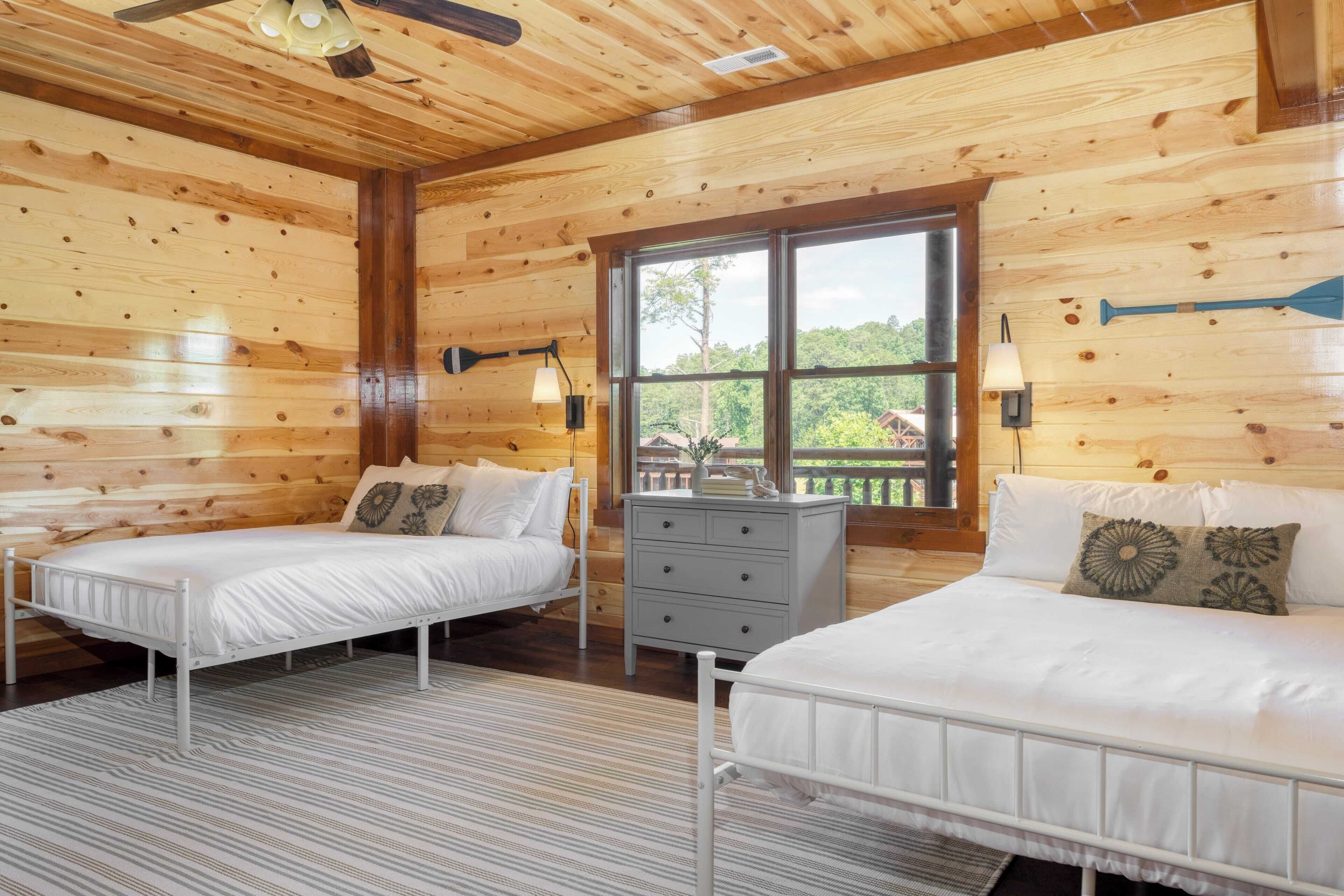 A wooden cabin bedroom with two beds, a dresser, and a window with a view of trees.