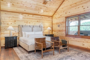 A wooden cabin bedroom with a bed, two chairs, a nightstand, a window, and a view of trees.