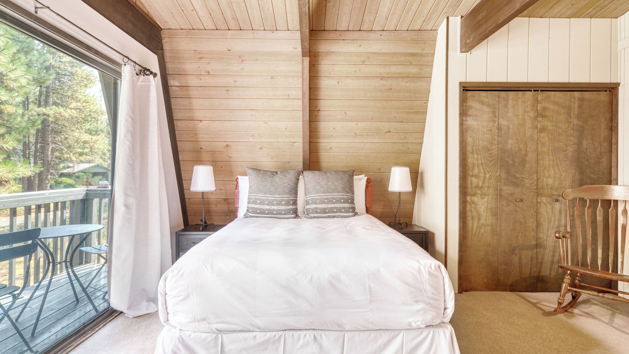 A wooden-paneled room with a bed, two bedside lamps, a wooden door, and a balcony with a table and chairs.