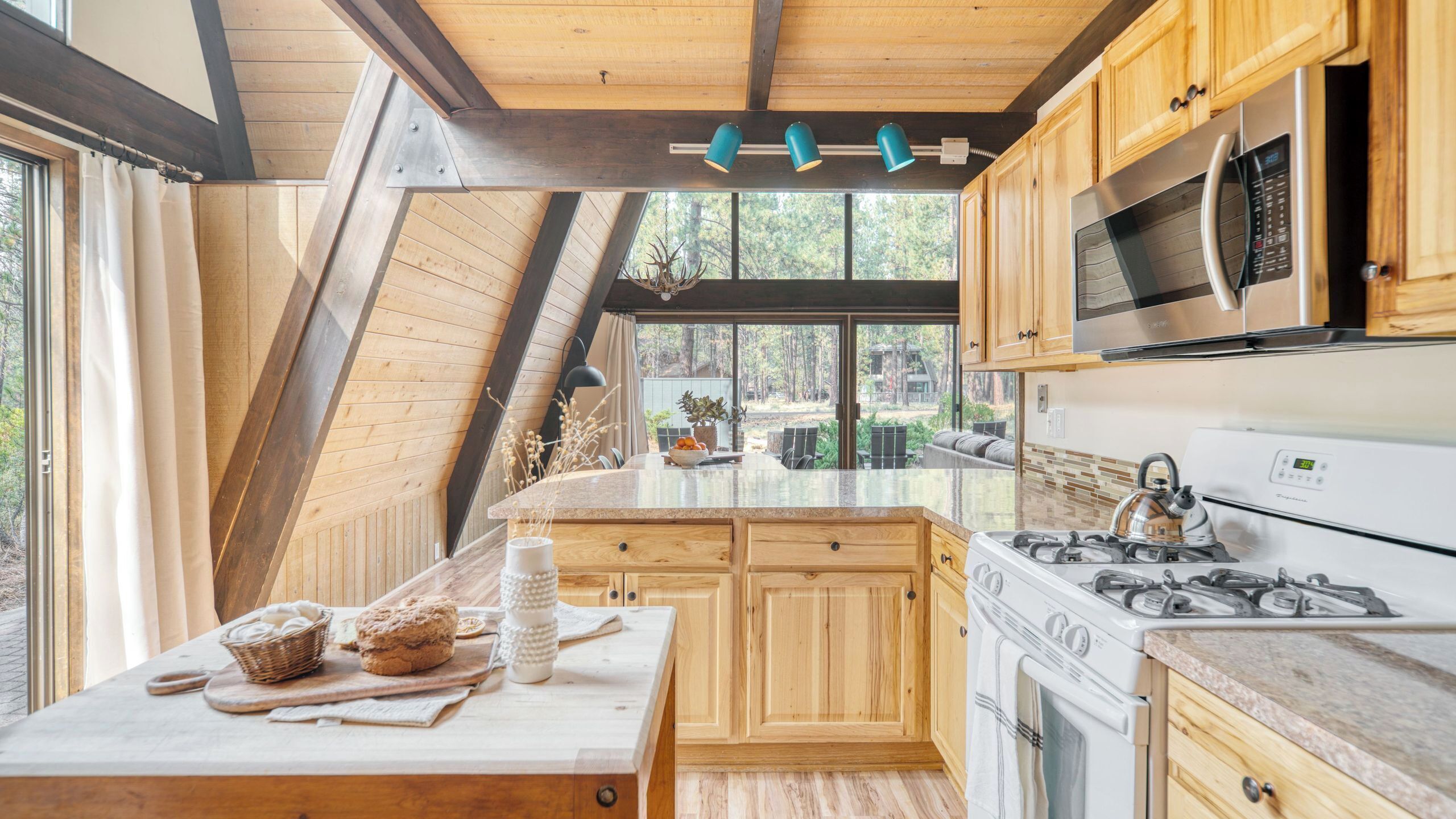 A kitchen with wooden cabinets, a gas stove, a microwave, and a large window overlooking a forest.