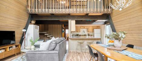 A spacious living area with a grey sofa, wooden flooring, a dining table with chairs, a kitchen area, and a balcony with a railing.