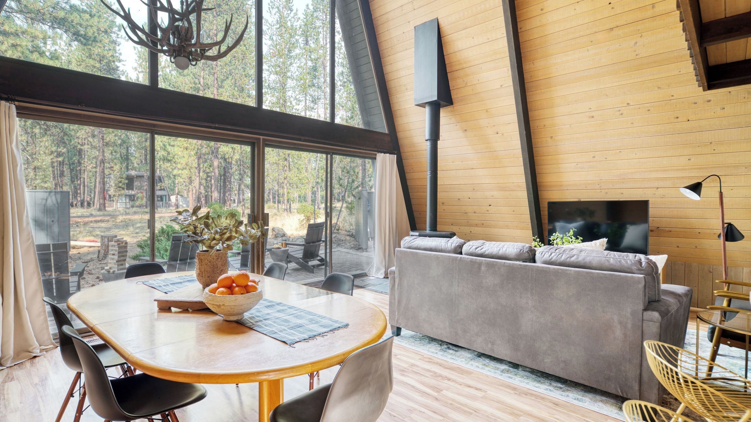A modern interior with a wooden ceiling, a sofa, a dining table with chairs, and a large window overlooking a forest.