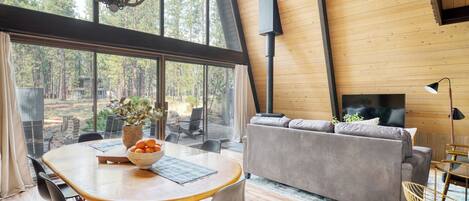 A modern interior with a wooden ceiling, a sofa, a dining table with chairs, and a large window overlooking a forest.