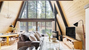 A cozy loft-style room with a large window, a sofa, a TV, and a fireplace. - Alder by Avantstay Hot Tub + Fireplace + Foosball Loft + Patio Grill Walk to Sunriver Village + Trails + Mt. Bachelor 30 Min (Bend)