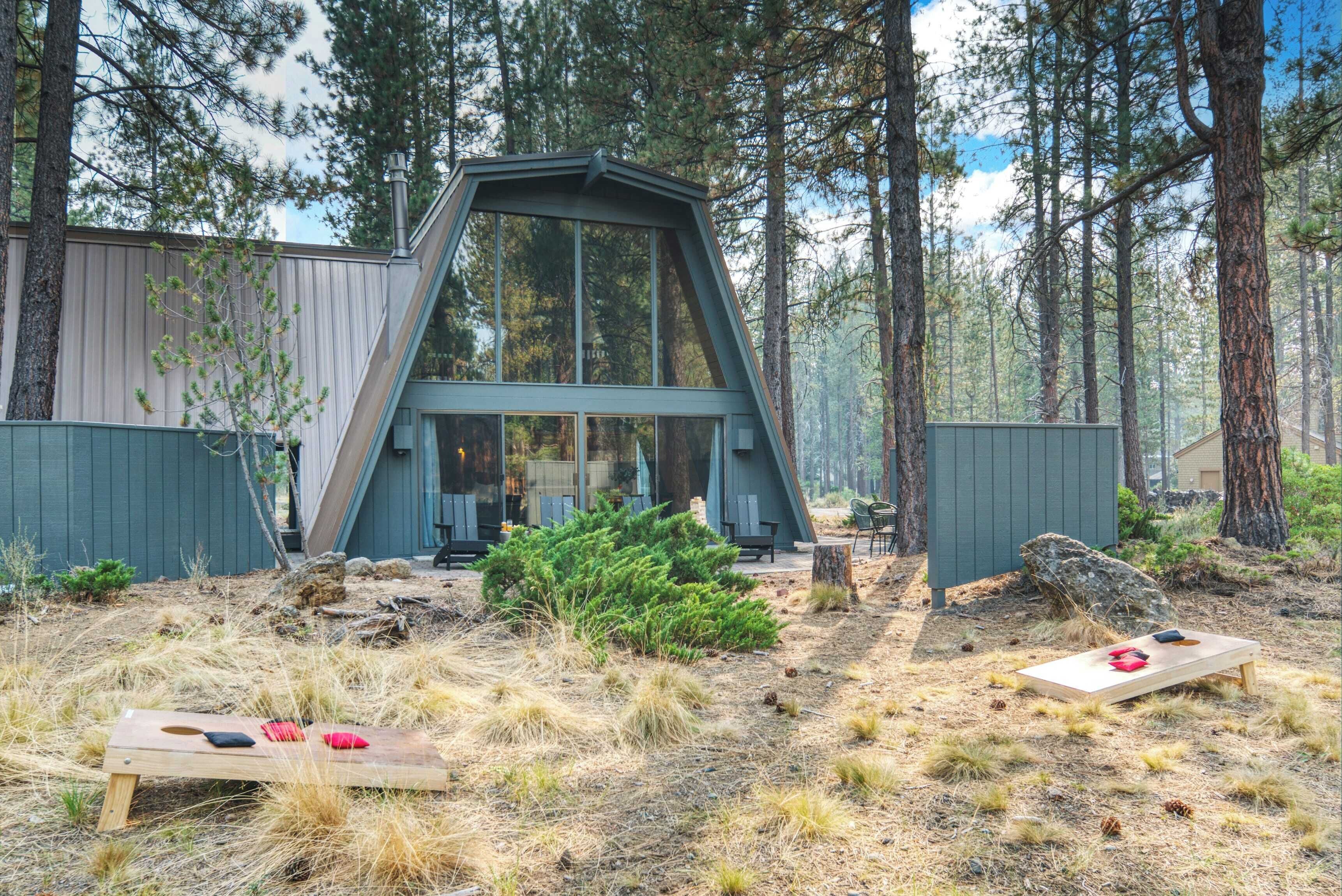 A modern cabin with a large glass window, surrounded by a forest. There are two wooden benches with items on them.