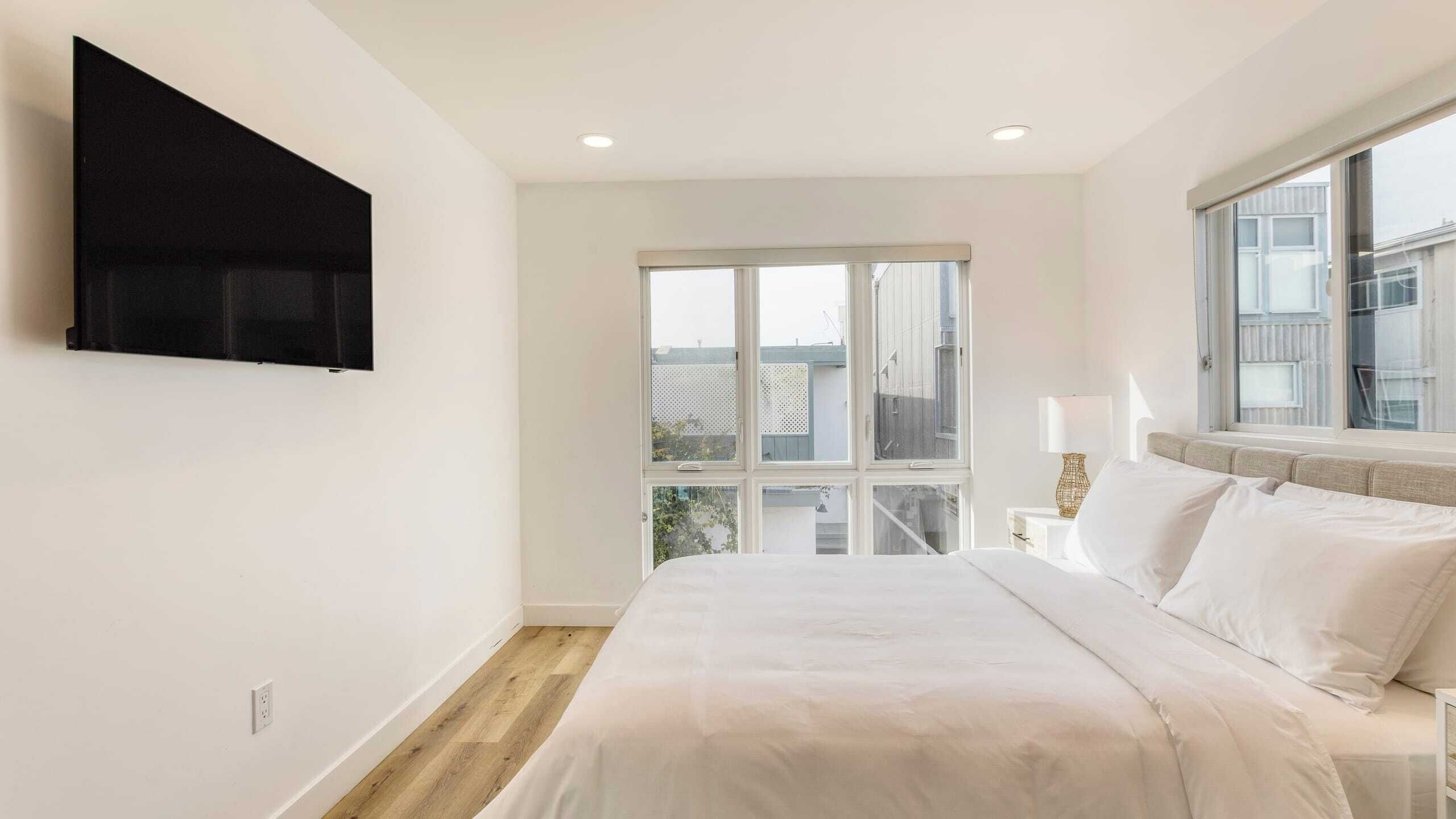 A modern bedroom with a large bed, a flat-screen TV mounted on the wall, and a large window offering a view of the city.