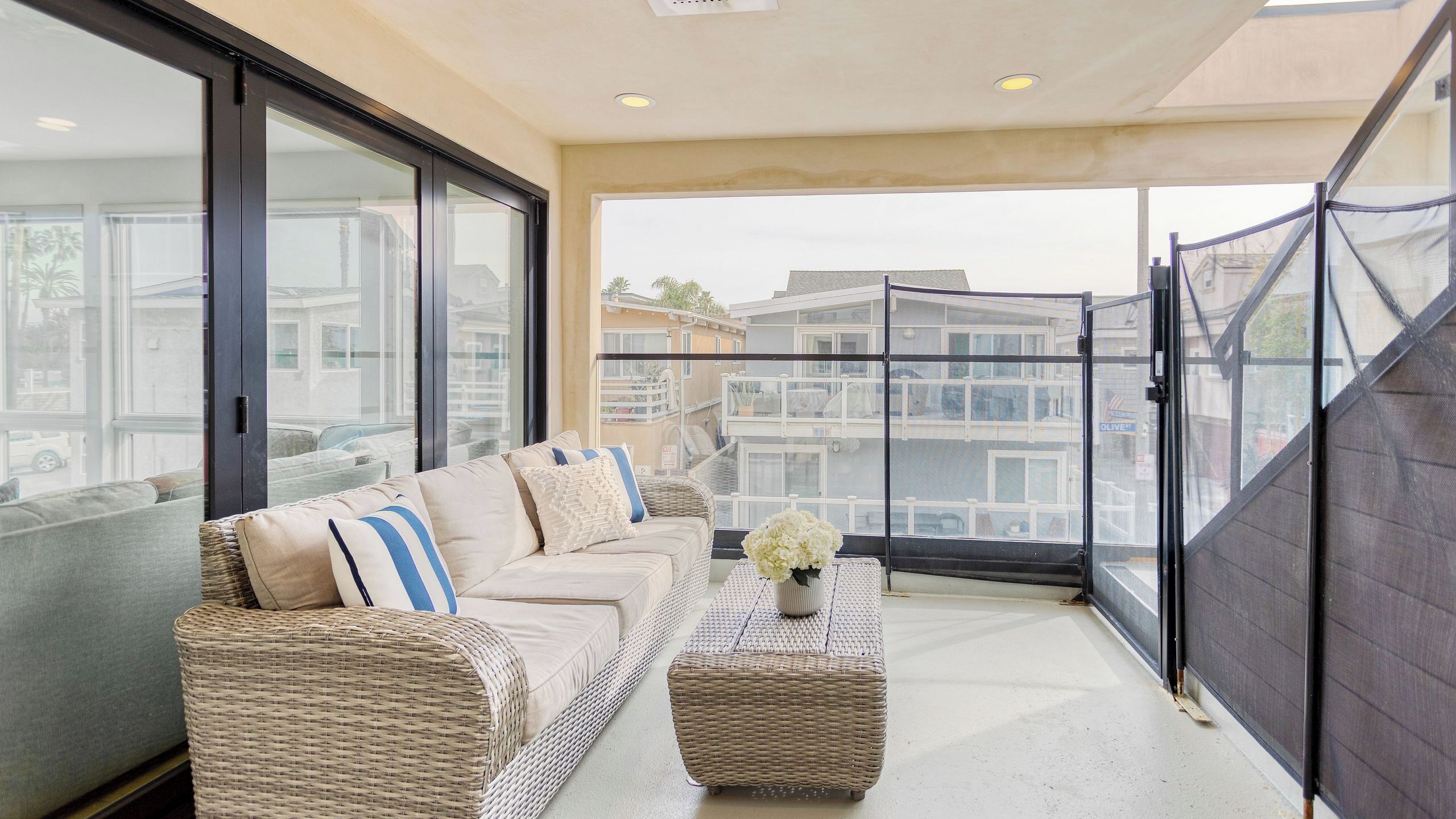 A balcony with a sofa, wicker ottoman, and a vase of flowers.