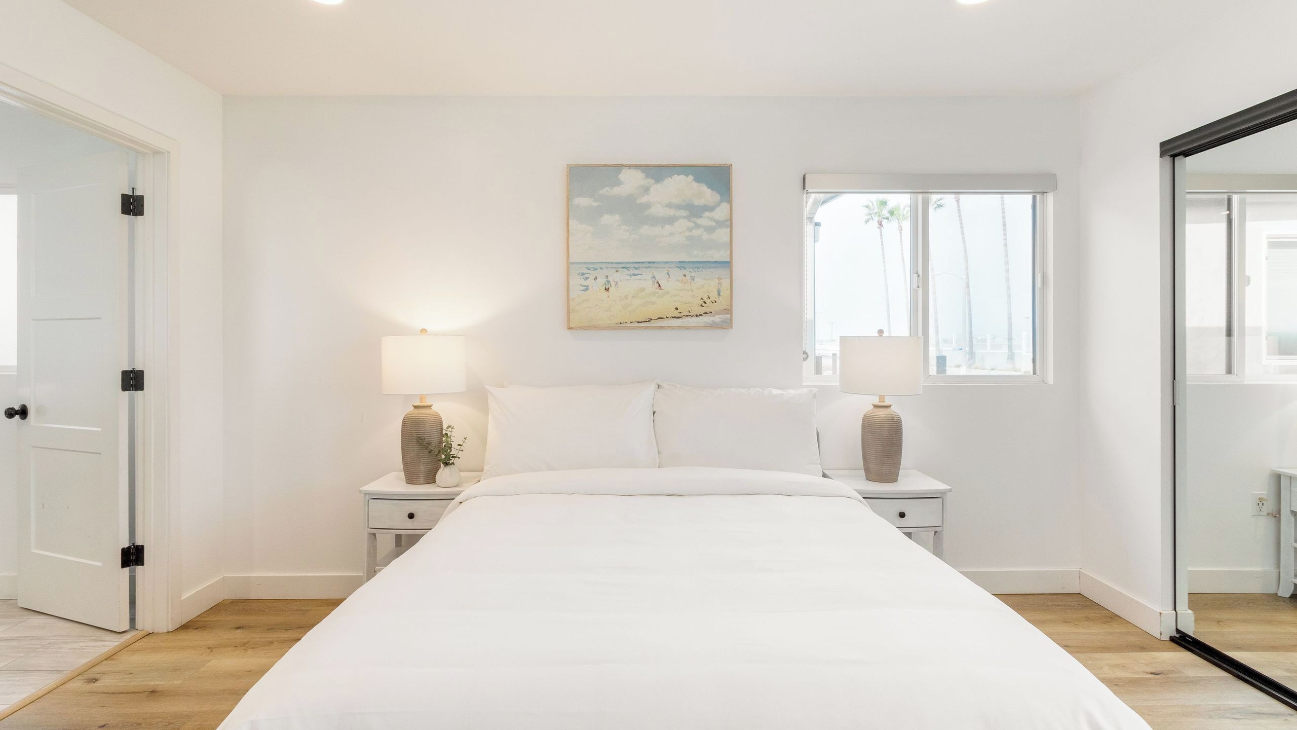 Um quarto com uma cama grande, duas mesas de cabeceira com luminárias, um quadro de uma praia e uma janela com vista para palmeiras.