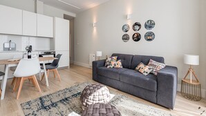 A modern living room with a grey sofa, patterned cushions, and a coffee table. There is a dining area with a wooden table and chairs, and a kitchen in the background.