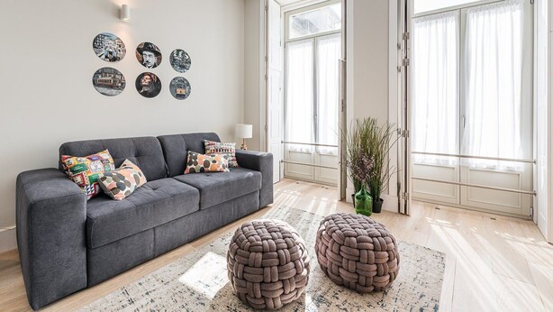 A modern living room with a grey sofa, patterned cushions, and a large window with sheer curtains.
