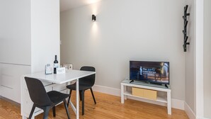 A modern, minimalistic room with a white table, two black chairs, a flat-screen TV on a white stand, and a wall-mounted shelf. - Sunny getaway in Rua do Bonjardim (Porto)