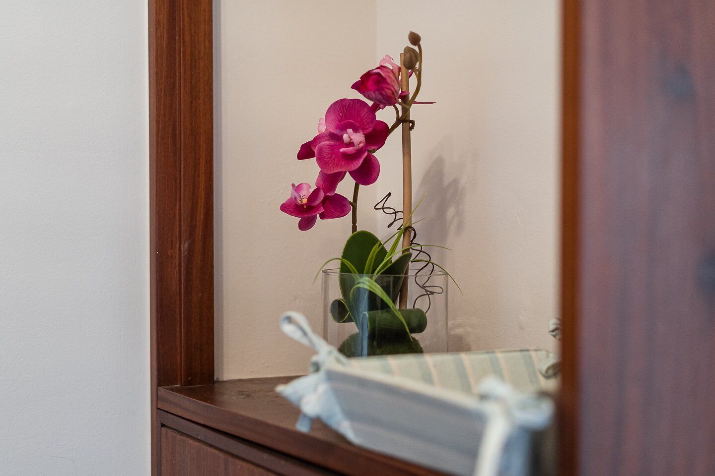 Un miroir reflétant une orchidée rose dans un vase en verre transparent posé sur une commode en bois.