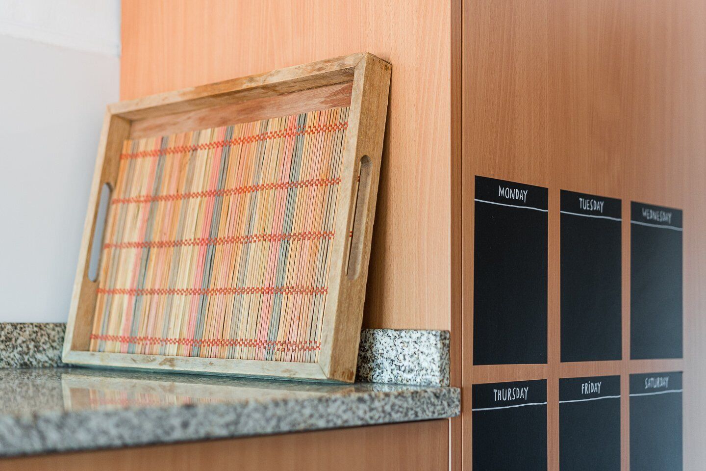 Un pannello di legno con motivo intrecciato, un calendario con lavagna e giorni della settimana, e un piano di lavoro in granito.