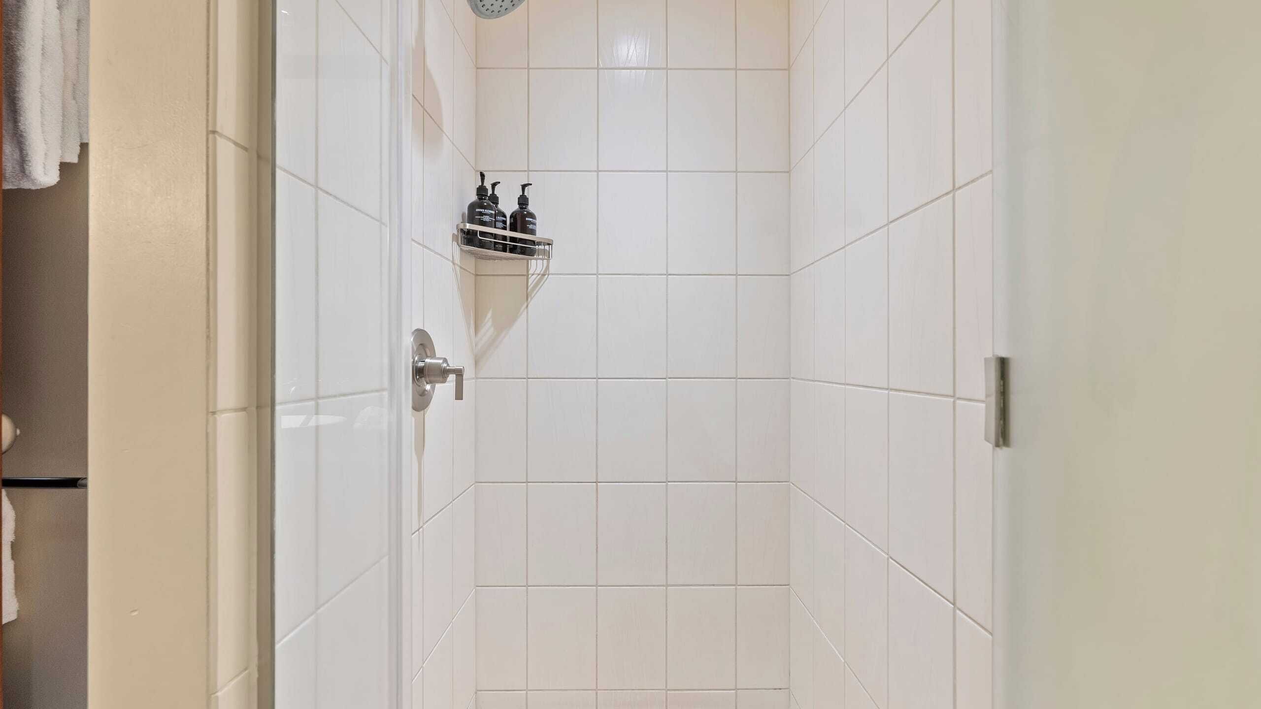A shower area with white tiles, a showerhead, and two bottles of shampoo and conditioner on a shelf.