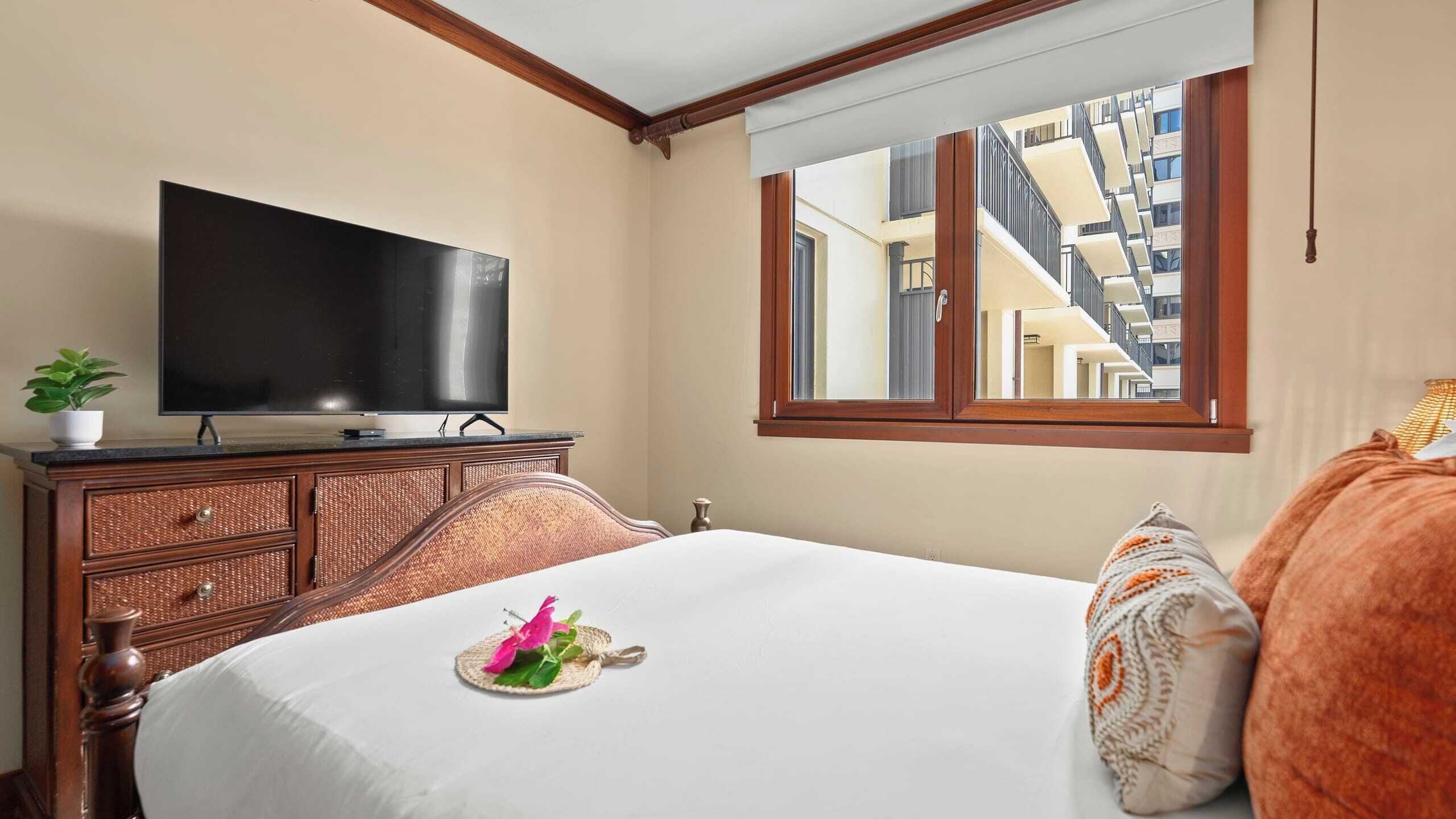 Habitación de hotel con cama, televisión sobre una cómoda de madera, ventana con vistas a edificios y una planta en la cómoda.