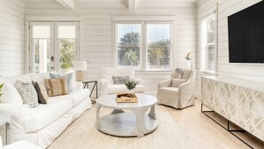 Un salon spacieux avec un canapé blanc, deux fauteuils, une table basse ronde et une grande fenêtre équipée de stores.