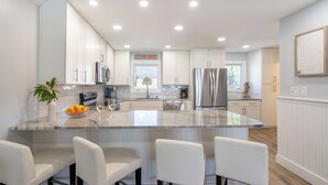 Una cocina moderna con armarios blancos, una isla central con asientos, electrodomésticos de acero inoxidable y un cartel que dice 'LET'S DO SUNSET'.