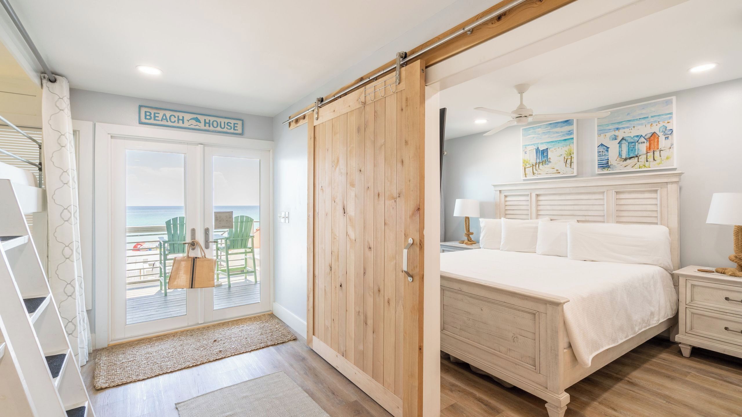 Ein Schlafzimmer mit einem Bett, Nachttischen, einem Holzschrank, einem Schild im Strandstil und Blick auf den Strand durch eine Tür.