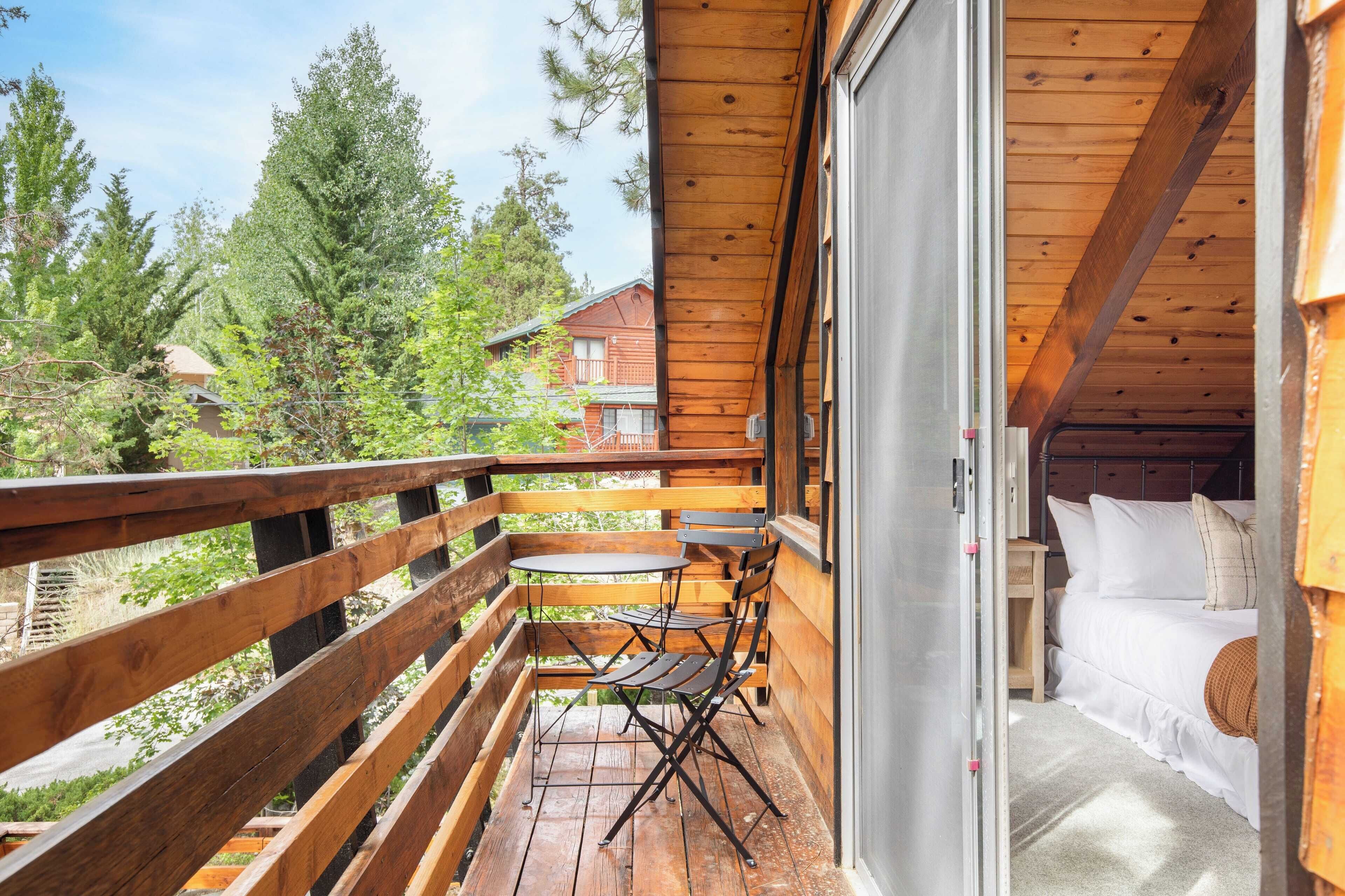 A wooden balcony with a table and chairs, a bed with white bedding, and a view of trees and buildings.