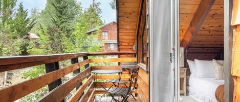A wooden balcony with a table and chairs, a bed with white bedding, and a view of trees and buildings.