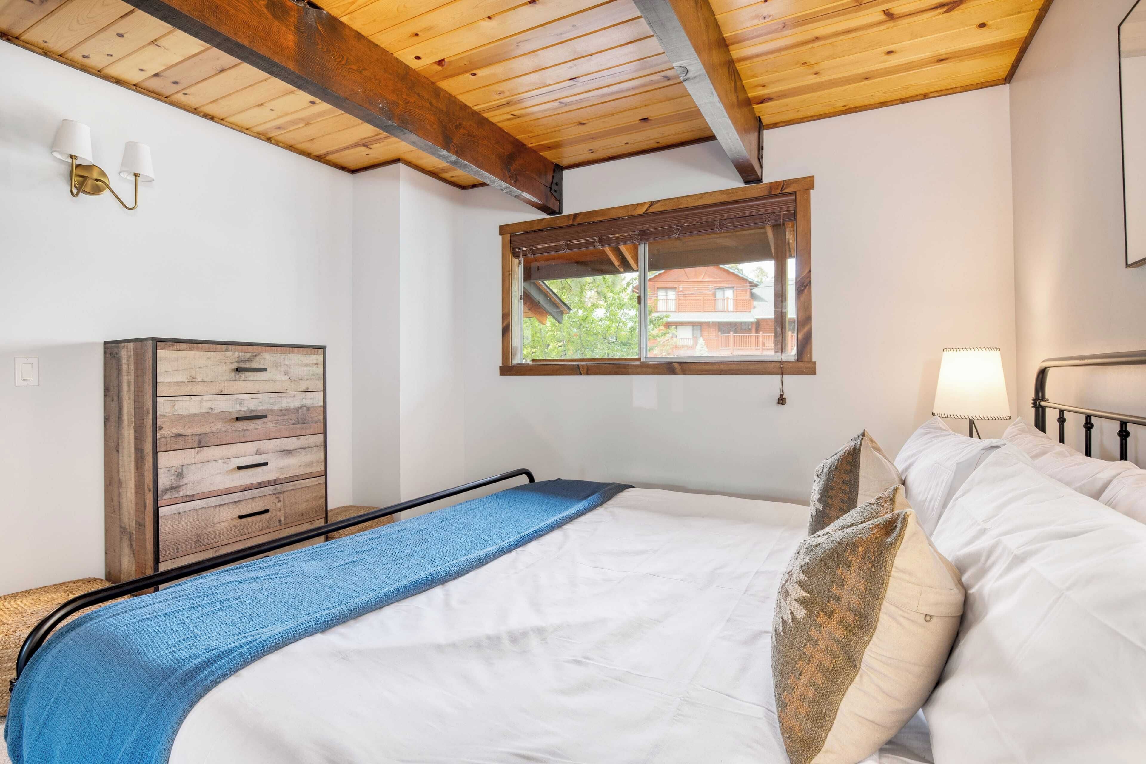 Une chambre avec un plafond en bois, une fenêtre donnant sur une maison, un lit avec une literie blanche, une couverture bleue et une commode en bois.
