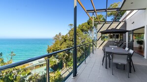 Een balkon met uitzicht op de oceaan, een eethoek met een tafel en stoelen, en een modern gebouw met een glazen deur.
