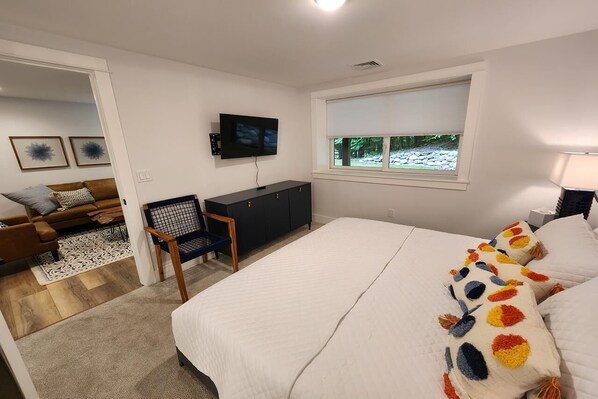 Een moderne slaapkamer met een groot bed, een aan de muur bevestigde televisie, een bureau en een raam met uitzicht op groen.
