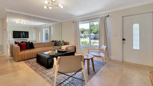 A modern living room with a large sofa, a coffee table, and a TV. There is a door with a glass panel and a view of the outside. - Summerfin by Avantstay Sunny Retreat Minutes From the Shore! (Pompano Beach)