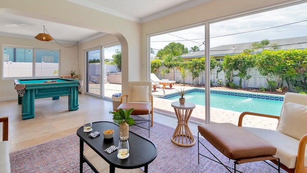 A spacious living room with a pool table, comfortable seating, and large windows overlooking a backyard. - Summerfin by Avantstay Sunny Retreat Minutes From the Shore! (Pompano Beach)