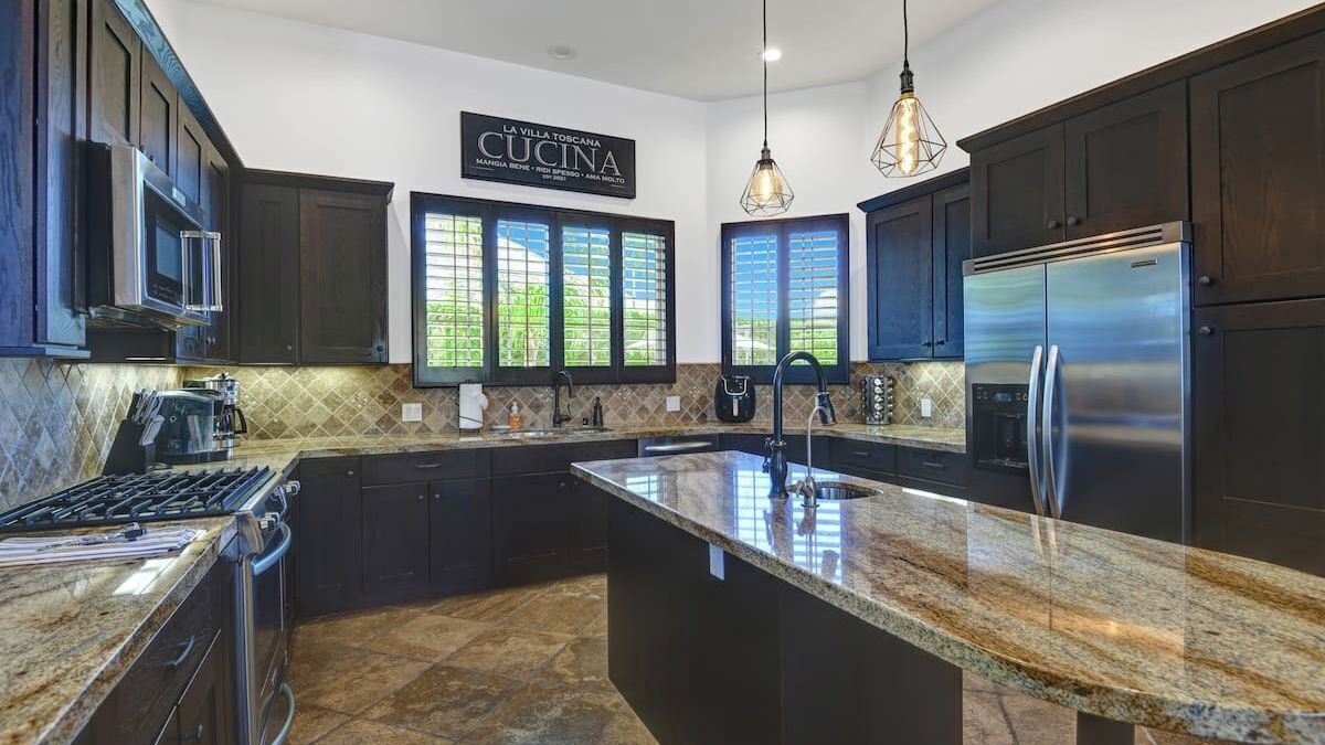 A modern kitchen with dark cabinetry, a stainless steel refrigerator, granite countertops, and pendant lighting.