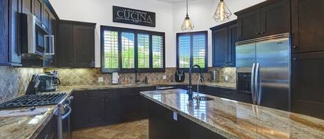 A modern kitchen with dark cabinetry, a stainless steel refrigerator, granite countertops, and pendant lighting.