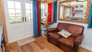 A room with a brown leather sofa, a wooden table with a lamp, a mirror with an ornate frame, and a view of a kitchen through a window with colorful curtains. - The Studio (York)