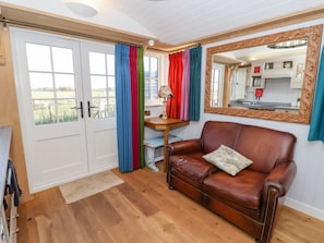 A room with a brown leather sofa, a wooden table with a lamp, a mirror with an ornate frame, and a view of a kitchen through a window with colorful curtains.