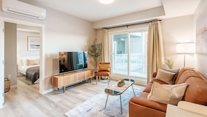 A modern living room with a sofa, a coffee table, a TV, and a large window with curtains. - Modern Condo w/AC, close to YYC (Calgary)