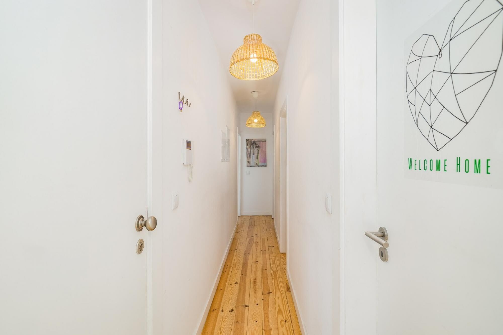 A hallway with wooden flooring, white walls, a hanging light fixture, and a 'WELCOME HOME' sign with a geometric design.