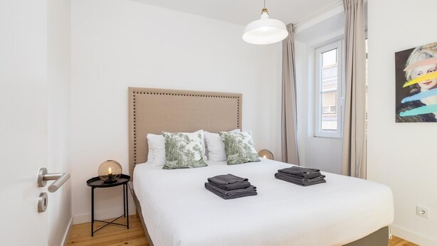 Un dormitorio ordenado con cama, mesitas de noche, una lámpara colgante y una ventana con cortinas.