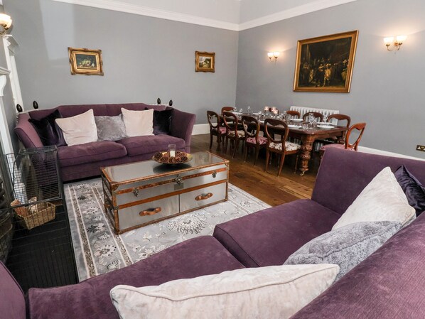 A living room with a purple sofa set, a glass coffee table with pinecones, and a dining area with wooden chairs and a table set for six. - The Stables and West Wing (York)