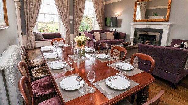 A dining room with a long wooden table set for six, flanked by purple upholstered chairs. A fireplace with a white mantel is visible in the background. - The Stables and West Wing (York)
