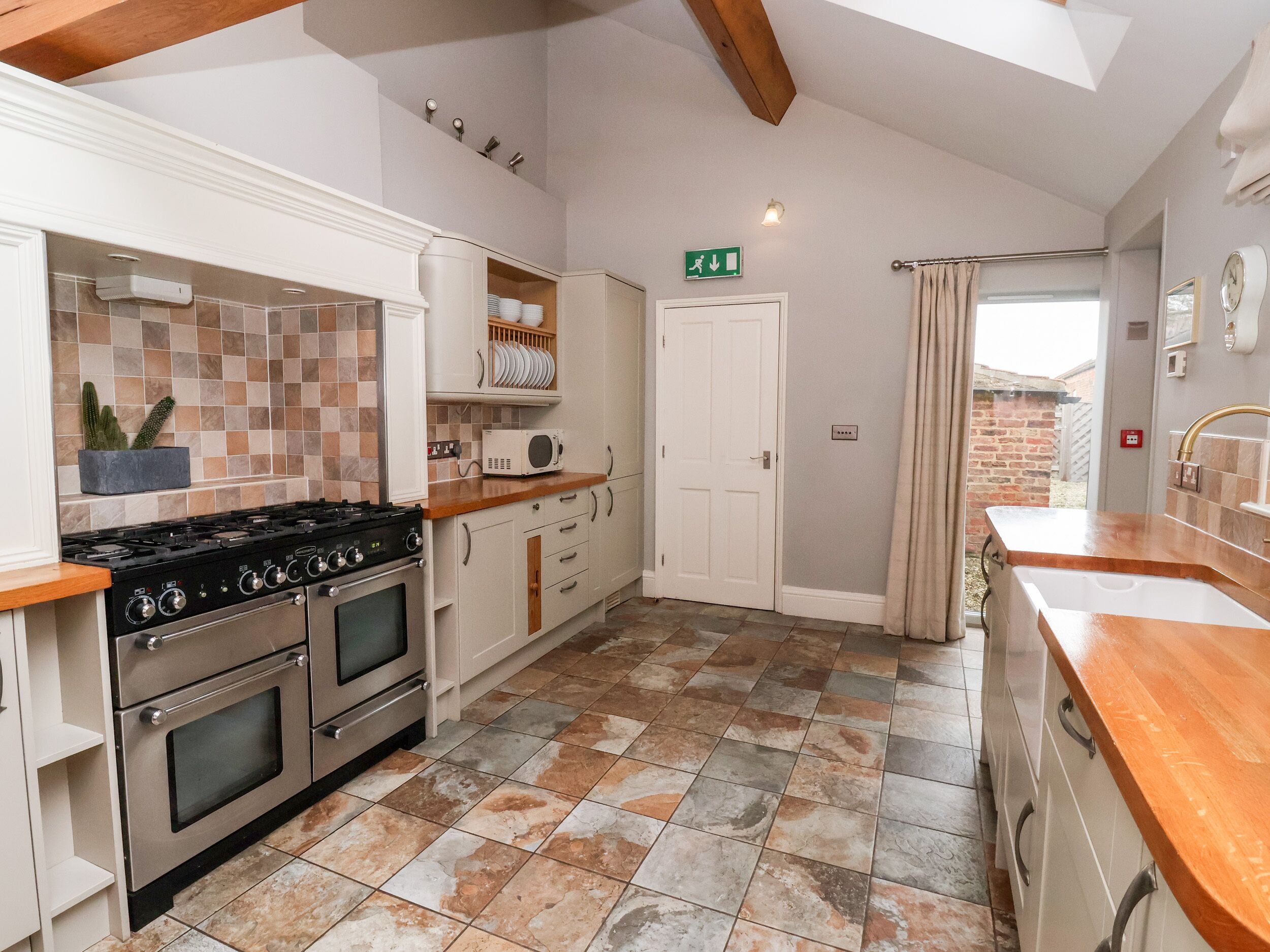 A kitchen with a gas stove, oven, microwave, and a sink. There is a door with an exit sign, and a window with curtains.