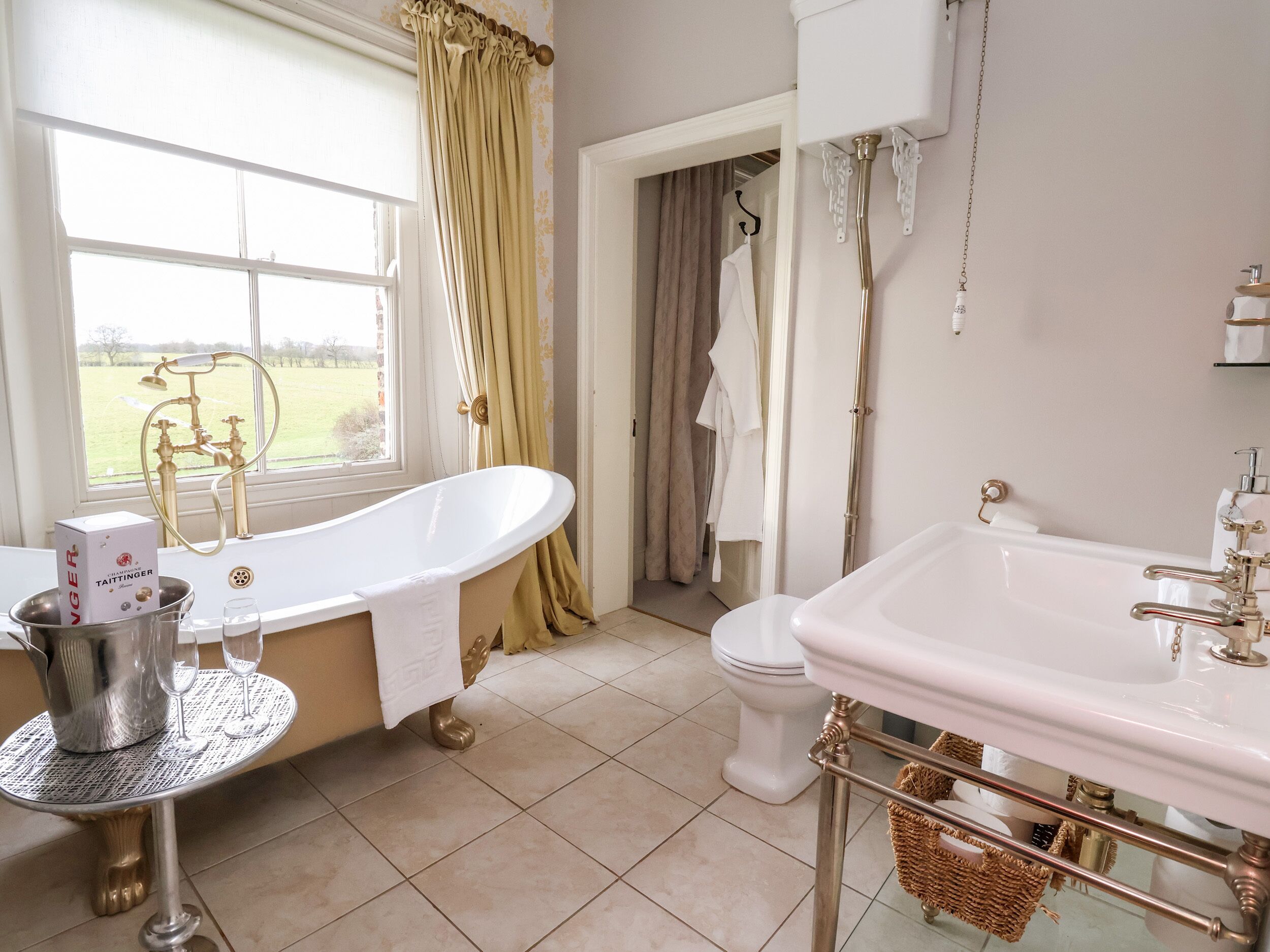A bathroom with a freestanding bathtub, a white sink with gold fixtures, and a window with curtains.