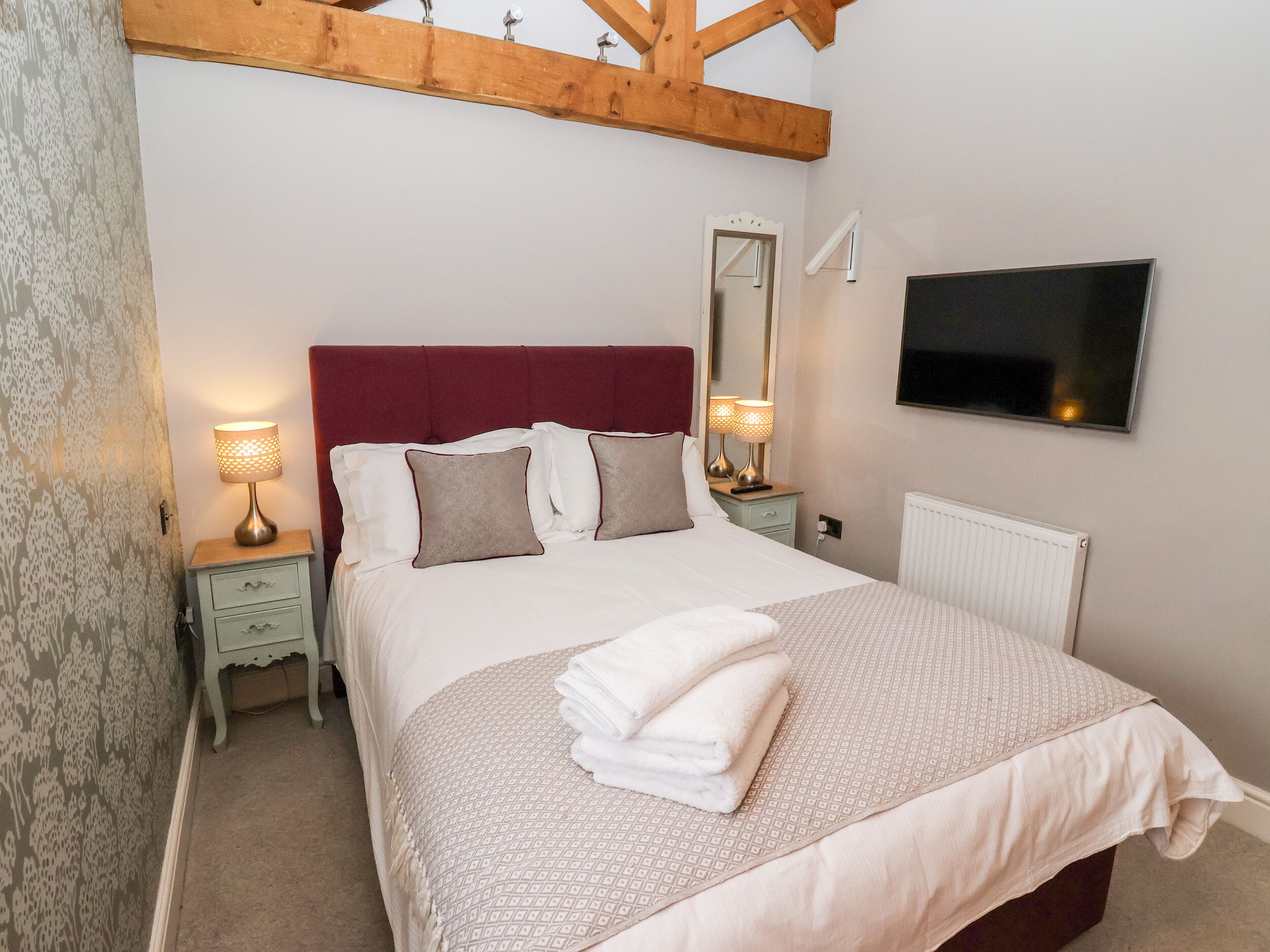 A neatly made bed with a red headboard, two bedside lamps, a TV mounted on the wall, and a radiator.
