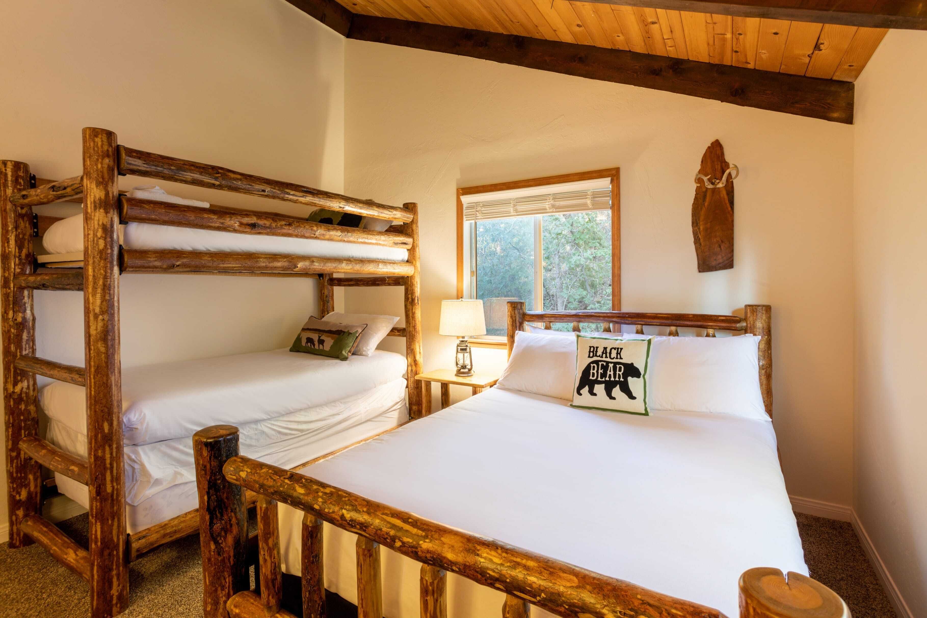 A rustic room with two bunk beds and a single bed, wooden beams on the ceiling, a window with a view of trees, and a wall-mounted artwork.