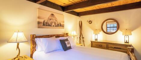 A bedroom with a wooden headboard, a bed with white linens, two bedside lamps, a framed picture on the wall, a wooden dresser, and a window with a view.