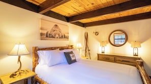 A bedroom with a wooden headboard, a bed with white linens, two bedside lamps, a framed picture on the wall, a wooden dresser, and a window with a view. - Bears Lair by Avantstay Wood-burning Fireplace + Firepit + Expansive Deck 3 Minutes to Bear Mountain + Big Bear Lake (Big Bear Lake)