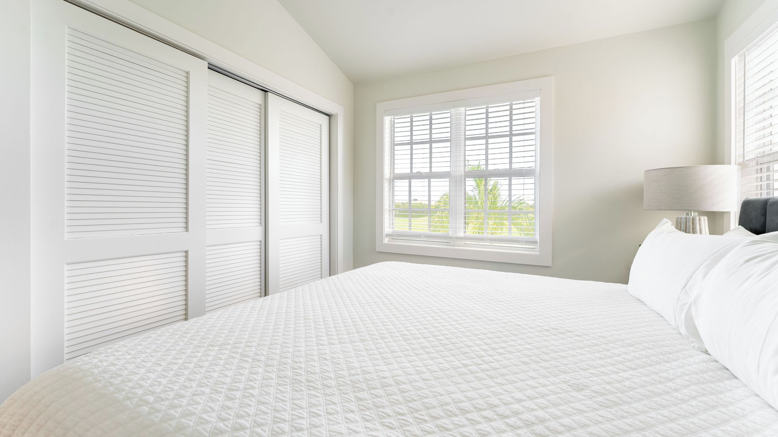 A bedroom with a large bed, a window with blinds, a nightstand with a lamp, and a closet with sliding doors.