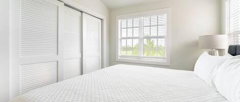 A bedroom with a large bed, a window with blinds, a nightstand with a lamp, and a closet with sliding doors.